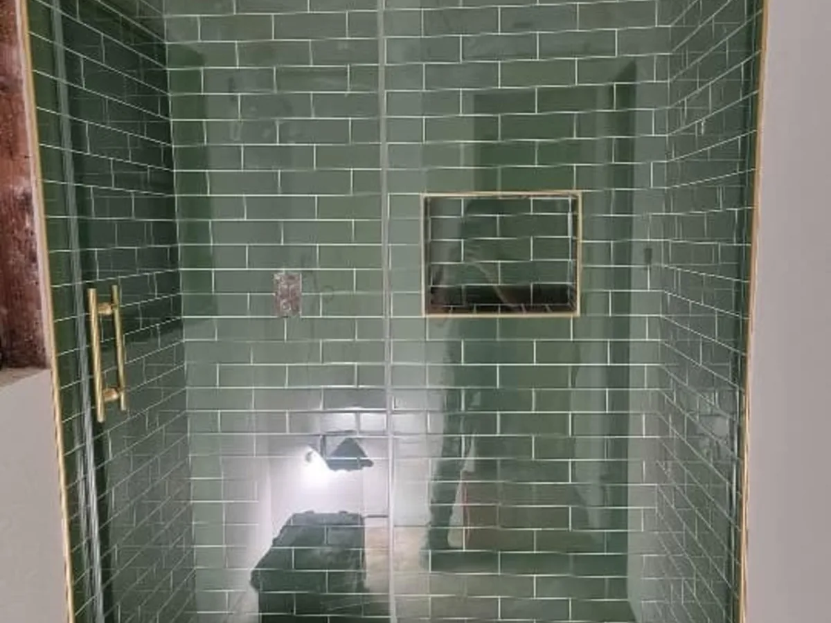Walk in shower lined with green subway tile and a glass door with gold trim